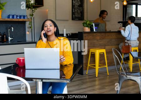 Woman wearing yellow sweater speaking on smartphone and working on laptop at cafe with red mug Stock Photo