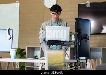 Hispanic male adjusting workstation at office sit-stand desk with laptop on monitor arm and screens Stock Photo