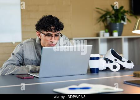 Man leaning over laptop at office desk, analyzing data next to VR headset, smartphone, coffee mug Stock Photo