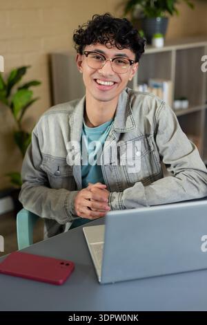 Man wearing denim jacket smiling at camera sitting at home office desk using laptop and smartphone Stock Photo