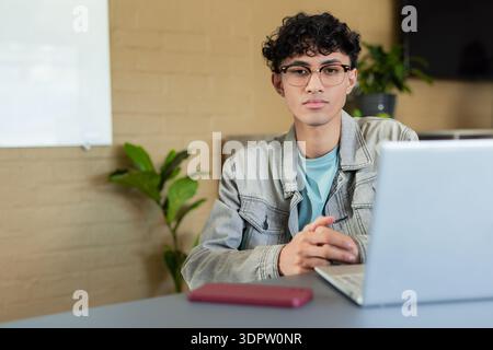 Asian man wearing denim jacket and eyeglasses sitting at office desk typing on laptop smartphone Stock Photo