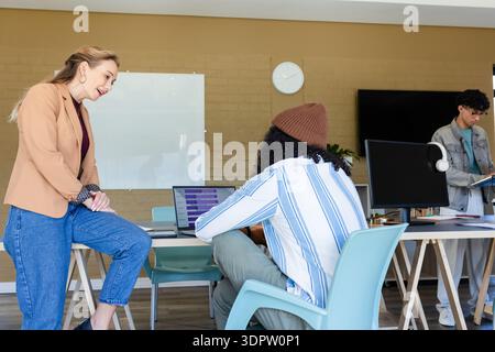 Diverse coworkers collaborating around communal desk in office using laptop, folder, whiteboard Stock Photo