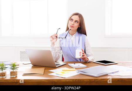 Woman Thinking While Holding Glasses at Desk With Laptop and Cup in Office Space Stock Photo