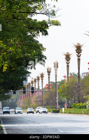 Wide urban road with palm-shaped street lamps, green trees lining pavement, cars driving under overcast sky in a modern city Stock Photo