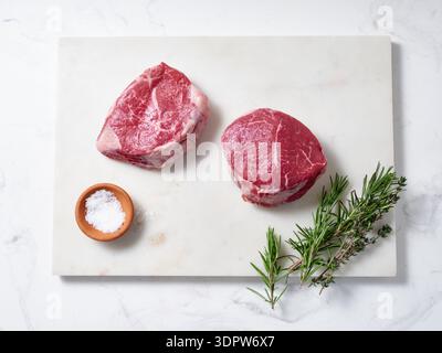 Two raw chuck tender steaks are placed on a marble cutting board. Salt, pepper, and fresh herbs are nearby. This scene shows ingredients for a healthy Stock Photo