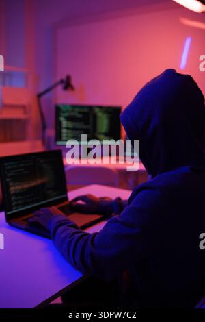 A mysterious hooded hacker working on a computer in a dark room. Stock Photo