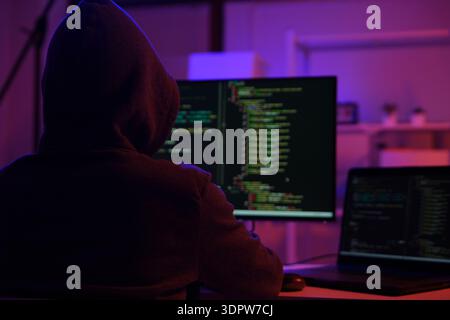 A mysterious hooded hacker working on a computer in a dark room. Stock Photo