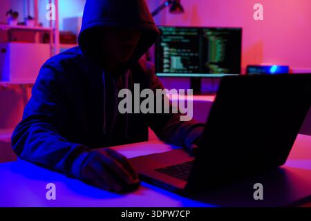 A mysterious hooded hacker working on a computer in a dark room. Stock Photo