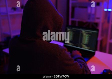 A mysterious hooded hacker working on a computer in a dark room. Stock Photo