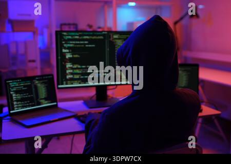 A mysterious hooded hacker working on a computer in a dark room. Stock Photo