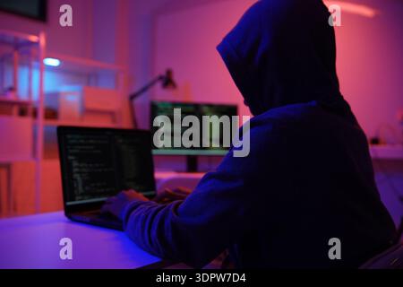 A mysterious hooded hacker working on a computer in a dark room. Stock Photo