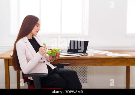 Working Woman Enjoys Salad at Desk While Focusing on Computer in Bright Office Setting Stock Photo