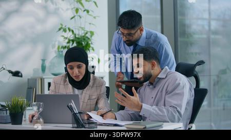 Business Arabian team three Indian colleagues in office discuss problem documents looking at computer happy celebrate work winning achieve looking at Stock Photo