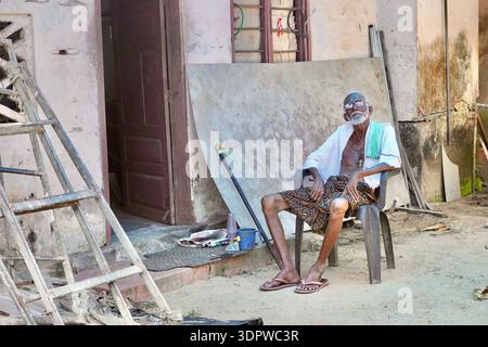Elderly Indian man resting on plastic chair outside weathered home in Alappuzha, Kerala, India Stock Photo