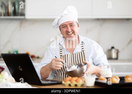 Man in chef hat and apron prepares cupcakes in kitchen according to recipe from the Internet - looking at laptop screen Stock Photo