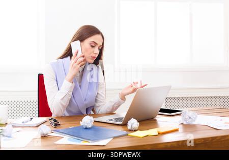 Worker Speaks on Phone While Using Laptop at Desk in Bright Office Space During Daytime Stock Photo