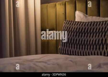 Close up of a modern hotel bed with white linen, patterned decorative pillow, and green upholstered headboard near curtains and wall outlet. Warm soft Stock Photo