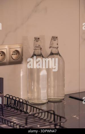 Two empty clear glass bottles with swing top caps standing on a kitchen countertop near an electrical outlet. Minimal domestic interior with soft warm Stock Photo