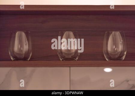 Three empty drinking glasses placed on a wooden shelf in a modern kitchen interior. Minimal composition with warm light and clean background creates a Stock Photo