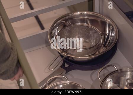Open kitchen drawer with neatly stored stainless steel cookware, including a metal pot and fine mesh strainer. Clean organized kitchen storage concept Stock Photo