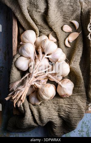 Wooden box filled with garlic bulbs with cloves placed on textured kitchen cloth. Raw organic vegetables in rustic food composition. Flat lay, top view Stock Photo