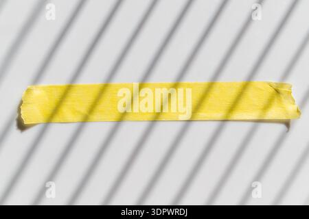 Torn strip of bright yellow masking tape on a light surface, with strong parallel shadows from window blinds creating a minimalist abstract compositio Stock Photo