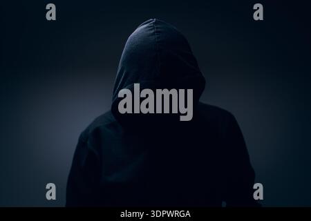 Dark hooded silhouette emerging from shadows, anonymous and enigmatic portrait symbolizing mystery, secrecy, tension, and the unknown. Selective focus Stock Photo