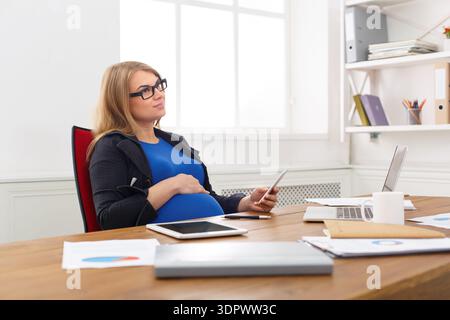 Woman Sitting in Office Holding Phone and Resting Hand on Pregnant Belly During Work Hours Stock Photo