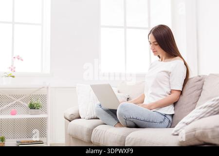 Woman Sitting on Couch Working on Laptop in Bright Living Room With Large Windows Stock Photo
