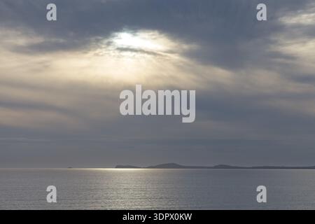 Sun rays breaking through dramatic clouds over a calm sea with distant islands on the Pembrokeshire coast, Wales. Stock Photo