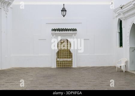 A wide-angle, minimalist view of a tranquil courtyard featuring a traditional tiled Moorish niche and a hanging vintage lantern centered on a vast whi Stock Photo