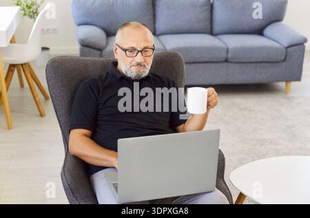 Senior man with laptop drinking coffee while working remotely in cozy home Stock Photo