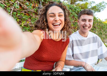 Young woman and transgender man laughing together on a sunny terrace during a casual outdoor gathering. Authentic lifestyle portrait representing frie Stock Photo