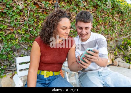 Young woman and transgender man sitting close together while checking a smartphone on an outdoor terrace. Candid lifestyle portrait representing frien Stock Photo