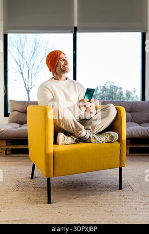 A cheerful man in a bright orange beanie and a white sweater sits cross-legged on a yellow armchair, holding a smartphone and smiling, with a cozy int Stock Photo