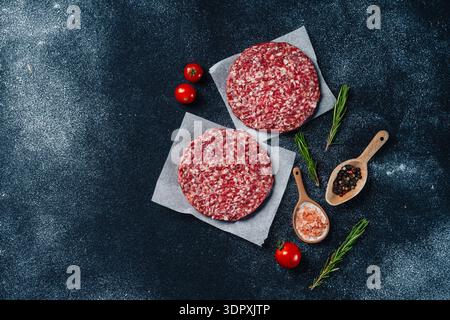 High-quality raw veal burger patties placed on parchment paper, accompanied by fresh rosemary sprigs, seasoning spices, and cherry tomatoes on a dark Stock Photo