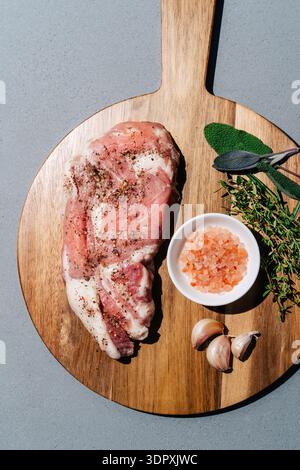 Raw ribeye steak seasoned with pepper and spices on a wooden board, accompanied by fresh herbs, garlic cloves, and bowl of pink salt, perfect for gour Stock Photo
