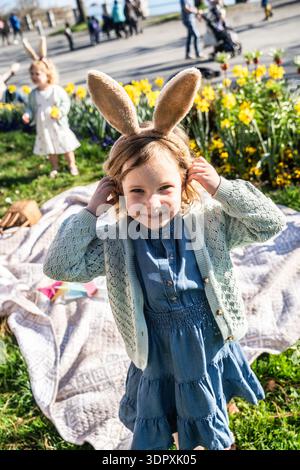 A joyful girl wearing bunny ears celebrates Easter outdoors with her family amidst blooming flowers. Sunny springtime creates a perfect setting for fe Stock Photo