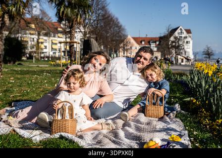 A cheerful family enjoys an Easter picnic outdoors during springtime. With the sun shining, parents and children are gathered on a blanket with Easter Stock Photo