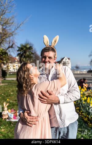 A couple joyfully celebrates Easter in a sunny spring park. They're embracing with smiles, surrounded by blooming flowers and festive decorations, cap Stock Photo