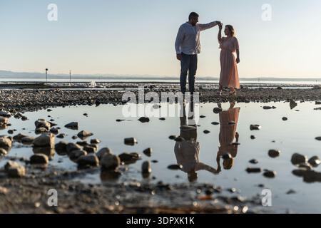 A couple enjoys a romantic moment by the seaside, dancing together on a rocky shore at sunset. Their reflection shimmers in the water, capturing a ser Stock Photo
