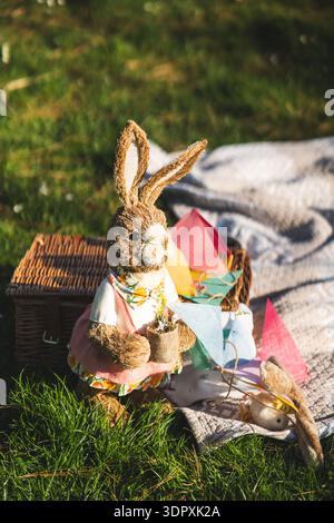A charming Easter bunny figure sits in the grass, surrounded by a picnic basket and colorful flags, capturing the joyful essence of springtime festivi Stock Photo
