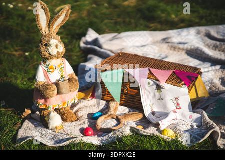 A charming spring picnic scene featuring an Easter bunny figure, colorful eggs, and a wicker basket on a blanket, creating a festive and playful Easte Stock Photo