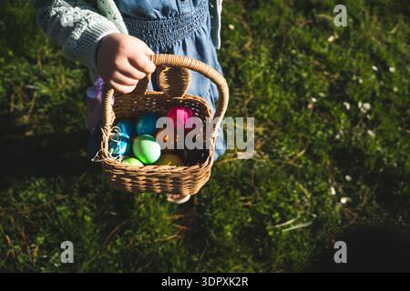 A girl celebrates Easter by holding a wicker basket filled with brightly colored eggs. The vivid hues of the eggs stand out against the lush green gra Stock Photo