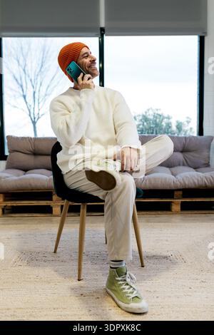 A man wearing a warm sweater and orange beanie sits cross-legged on a chair, smiling as he talks on his smartphone. The comfortable setting includes n Stock Photo