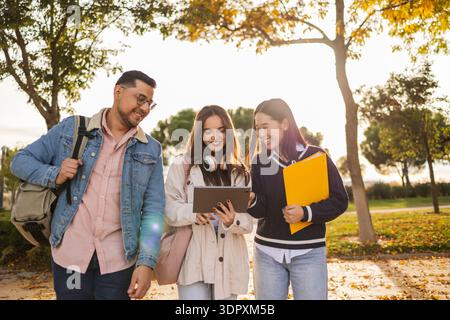 A group of three diverse college students walk together on a picturesque campus path, engaging with a tablet and carrying books, enjoying a vibrant au Stock Photo