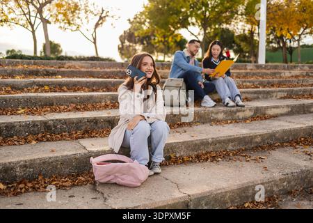 A group of students relaxes on stone steps, enjoying leisure time outdoors. One student smiles while talking on a phone, while two others review notes Stock Photo