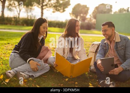 A group of three smiling students sit together on the grass in a sunlit park, enjoying a study session. They share resources like a tablet and a yello Stock Photo