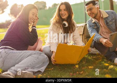 A group of three smiling students sit together on the grass in a sunlit park, enjoying a study session. They share resources like a tablet and a yello Stock Photo