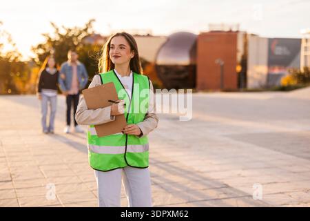 A student wearing a bright green safety vest holds a clipboard on a university campus. She appears focused and engaged, possibly working on a project. Stock Photo